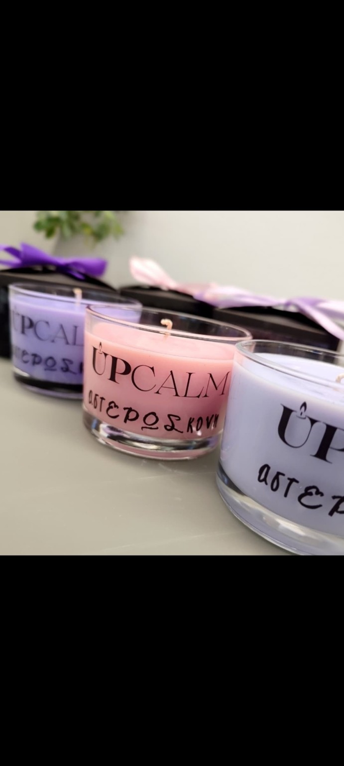 Ροζ και λιλά κεριά UPCALM σε διάφανα δοχεία με τον τίτλο Αστερόσκονη, δίπλα σε συσκευασίες με σατέν κορδέλες