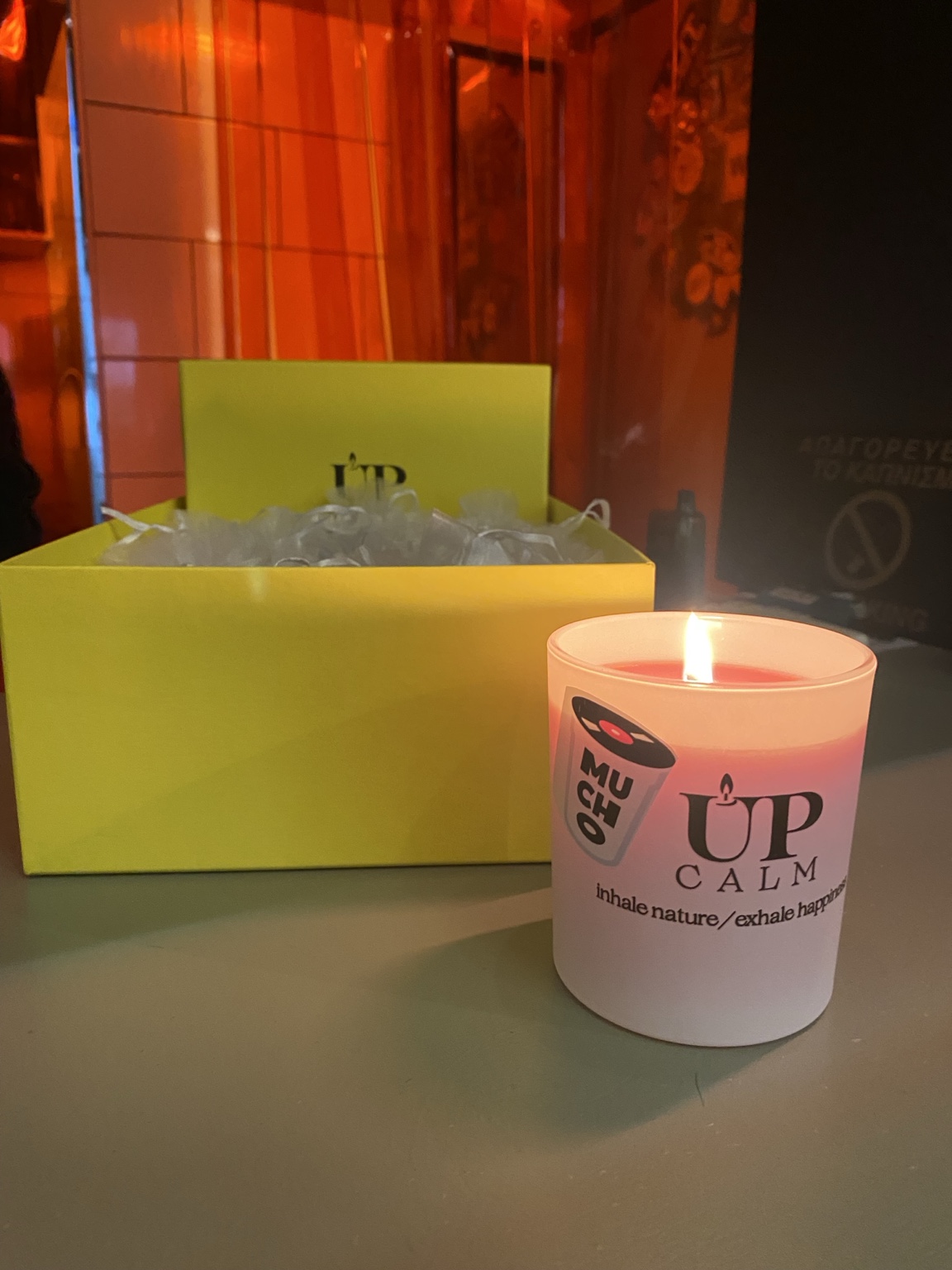 Κερί με λογότυπα Mucho και UPCALM, αναμμένο δίπλα σε κίτρινο κουτί δώρων στο opening night του Mucho Bar